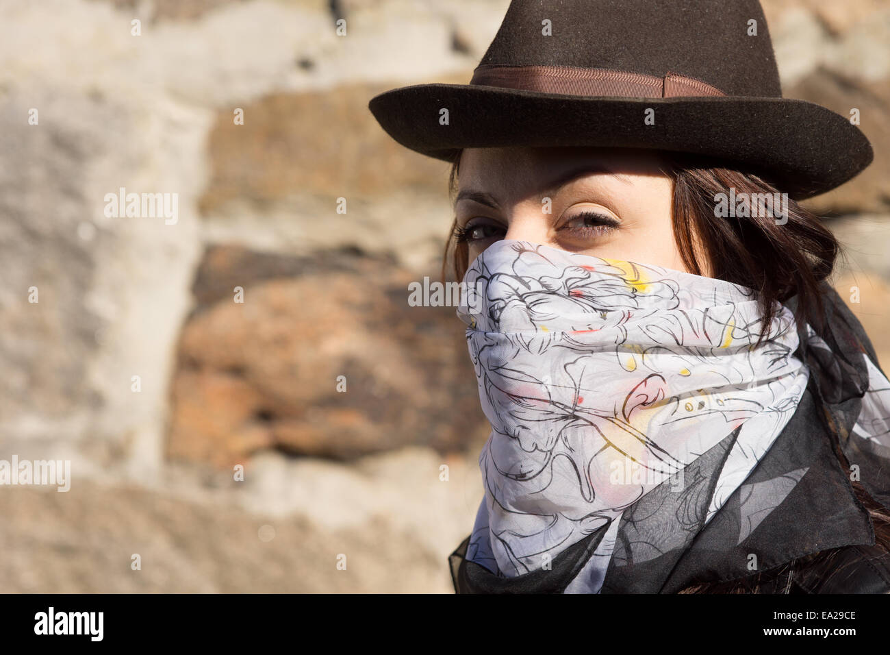 Trendige junge Frau trägt einen Schal über ihr Gesicht wie ein Bandit und ein stilvolles Hut Blick seitlich auf die Kamera gegen einen Stein Stockfoto