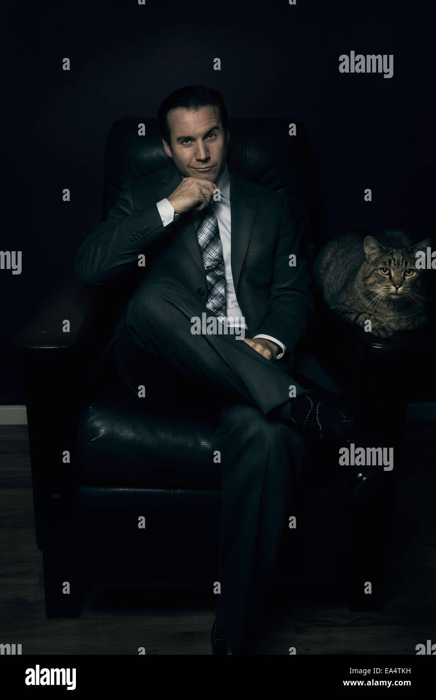Ein Mann in einem Anzug sitzt auf einem Stuhl mit einer Katze auf der Armlehne. Stockfoto