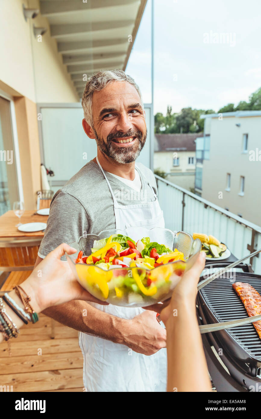 Porträt von lächelnden Mann auf seinem Balkon grillen, mit Frauenhand Salatschüssel im Vordergrund halten Stockfoto