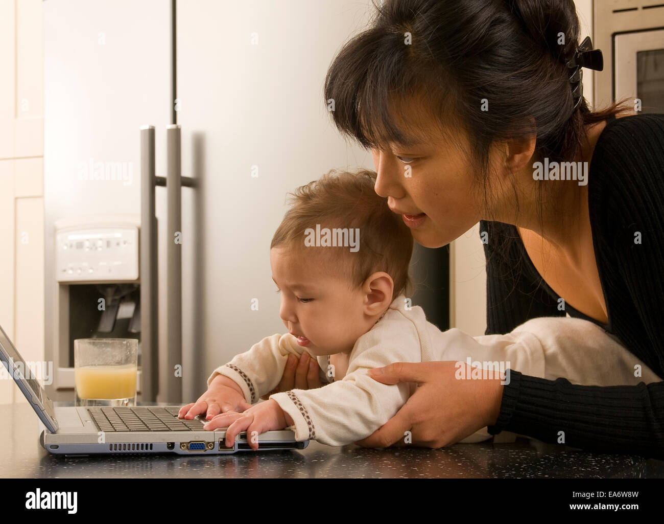 beschäftigt junge koreanische asiatische Mama mit Laptop-Computer in der modernen Küche hält 5 Monate altes gemischt Rennen (asiatische / caucasian) baby Stockfoto