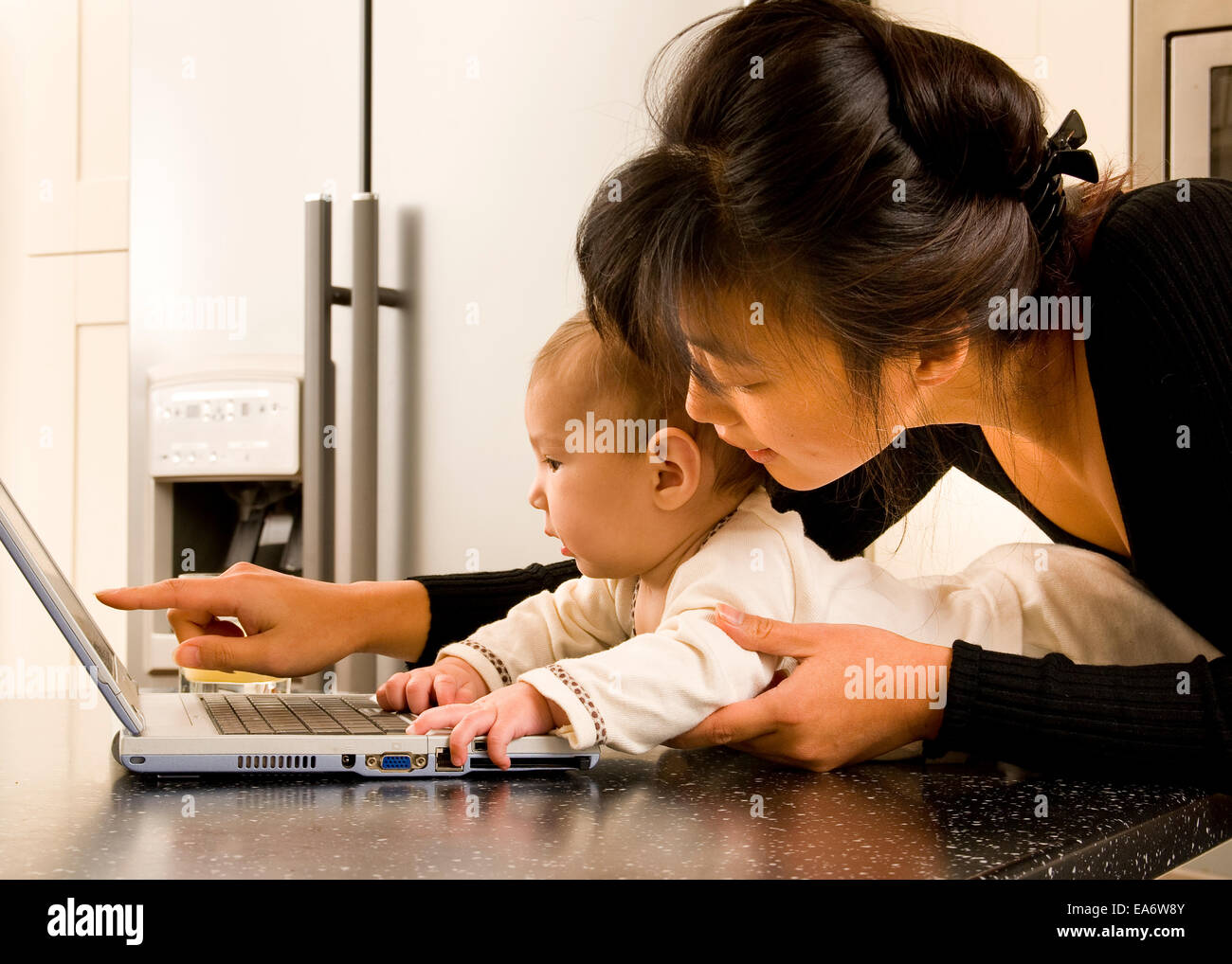 beschäftigt junge koreanische asiatische Mama mit Laptop-Computer in der modernen Küche hält 5 Monate altes gemischt Rennen (asiatische / caucasian) baby Stockfoto