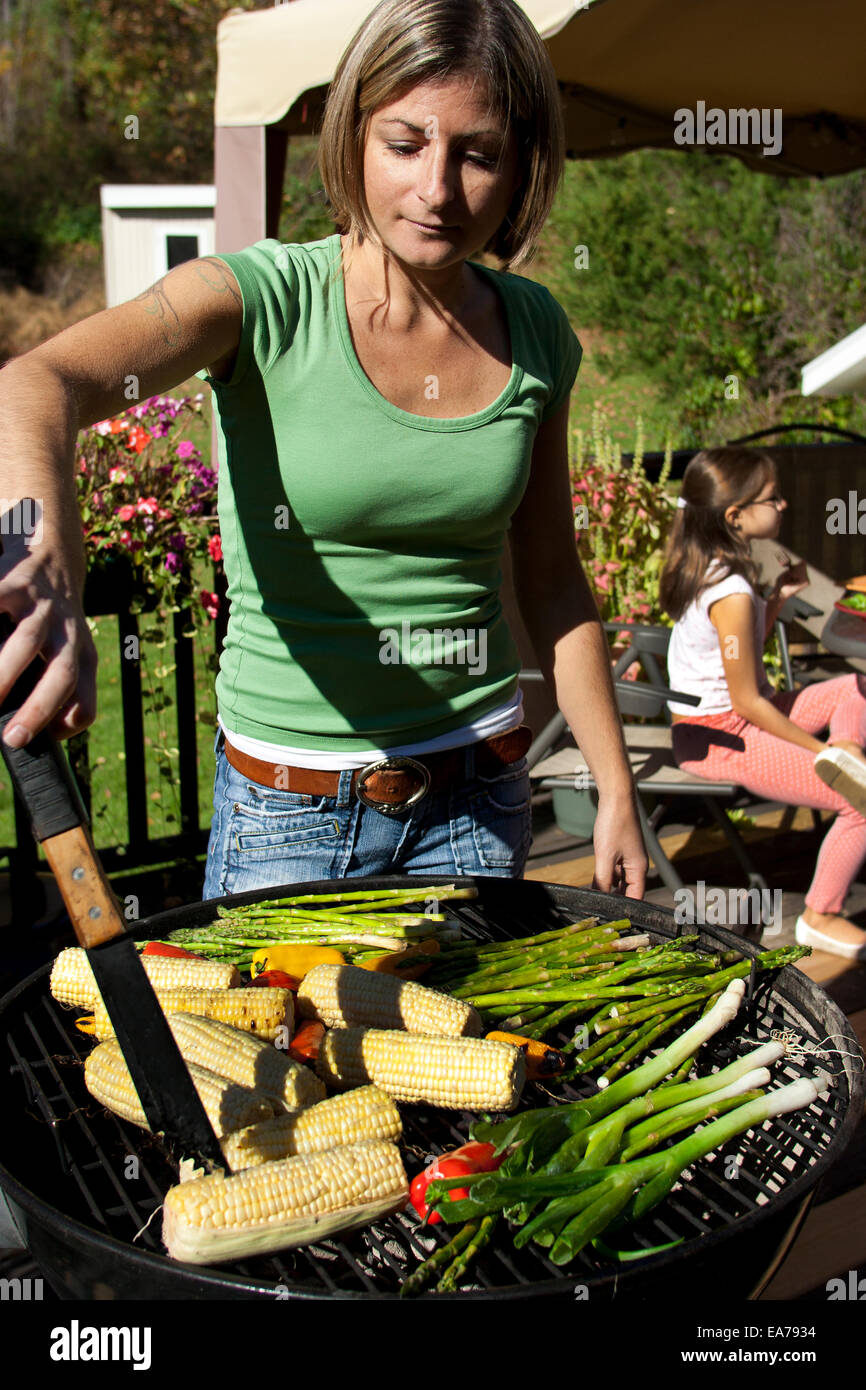 Mutter stand Barbecue-Grill mit Zangen mit Tochter (8-9) im Hintergrund Stockfoto
