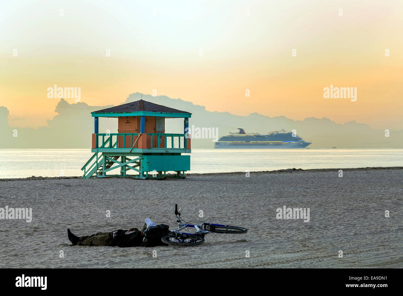 Mann schläft neben seinem Fahrrad an Miamis South Beach im Morgengrauen mit einem Bademeister Stand und ein Kreuzfahrtschiff im Hintergrund. Stockfoto