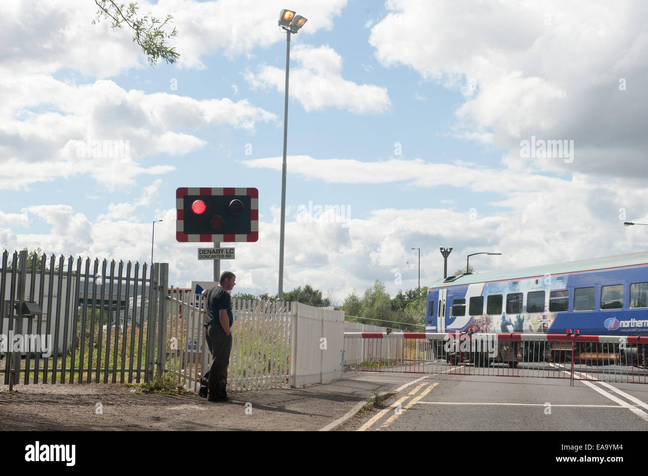 Der Zug fährt durch einen Bahnübergang, während ein Fußgänger hinter den Schranken wartet, während die Warnleuchten unter hellem, bewölktem Himmel blinken. Stockfoto
