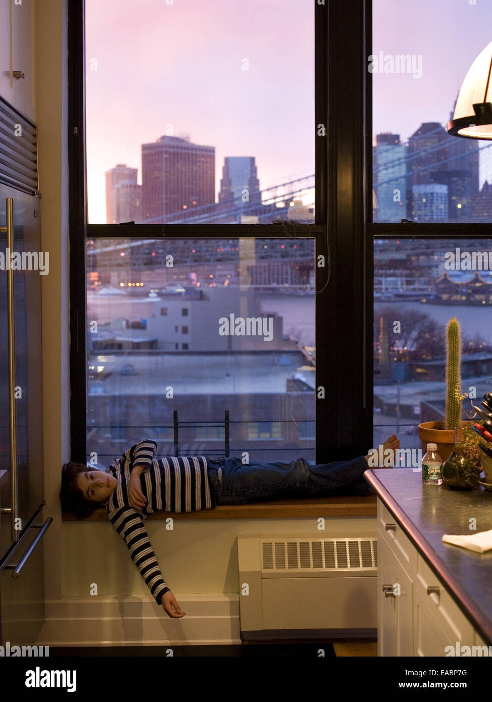 ein kleiner Junge liegend durch ein Fenster der Wohnung in der Stadt in der Abenddämmerung Stockfoto