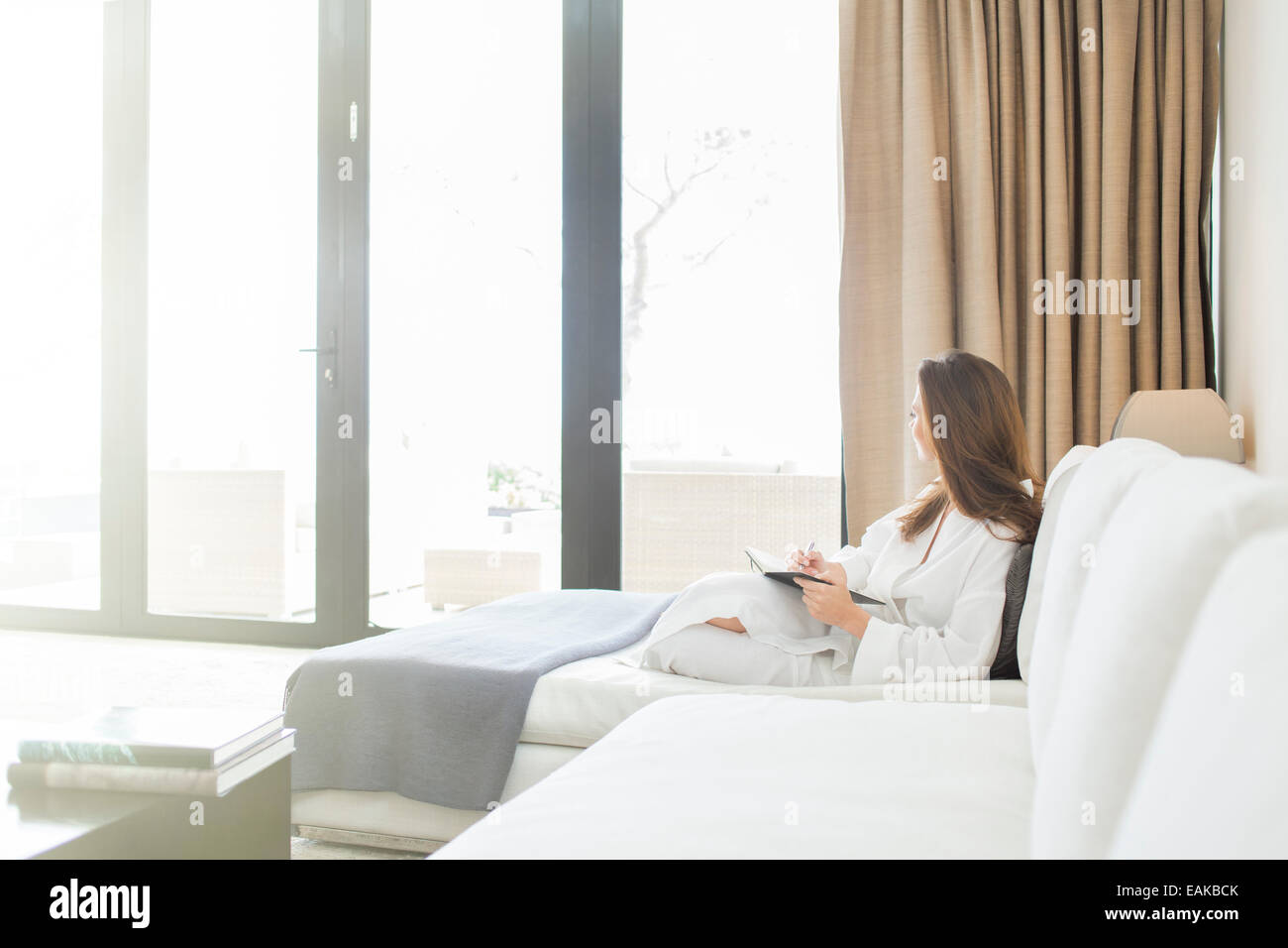 Frau trägt weißen Bademantel liegend auf dem Sofa im Wohnzimmer und Blick durch Fenster Stockfoto