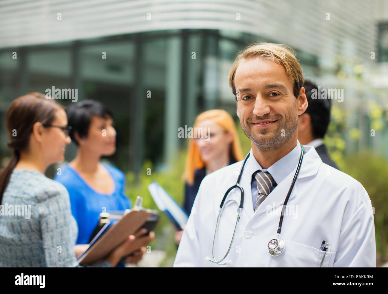 Porträt von lächelnden Arzt tragen Laborkittel vor Krankenhaus, Frauen mit Ablagen im Hintergrund stehend Stockfoto