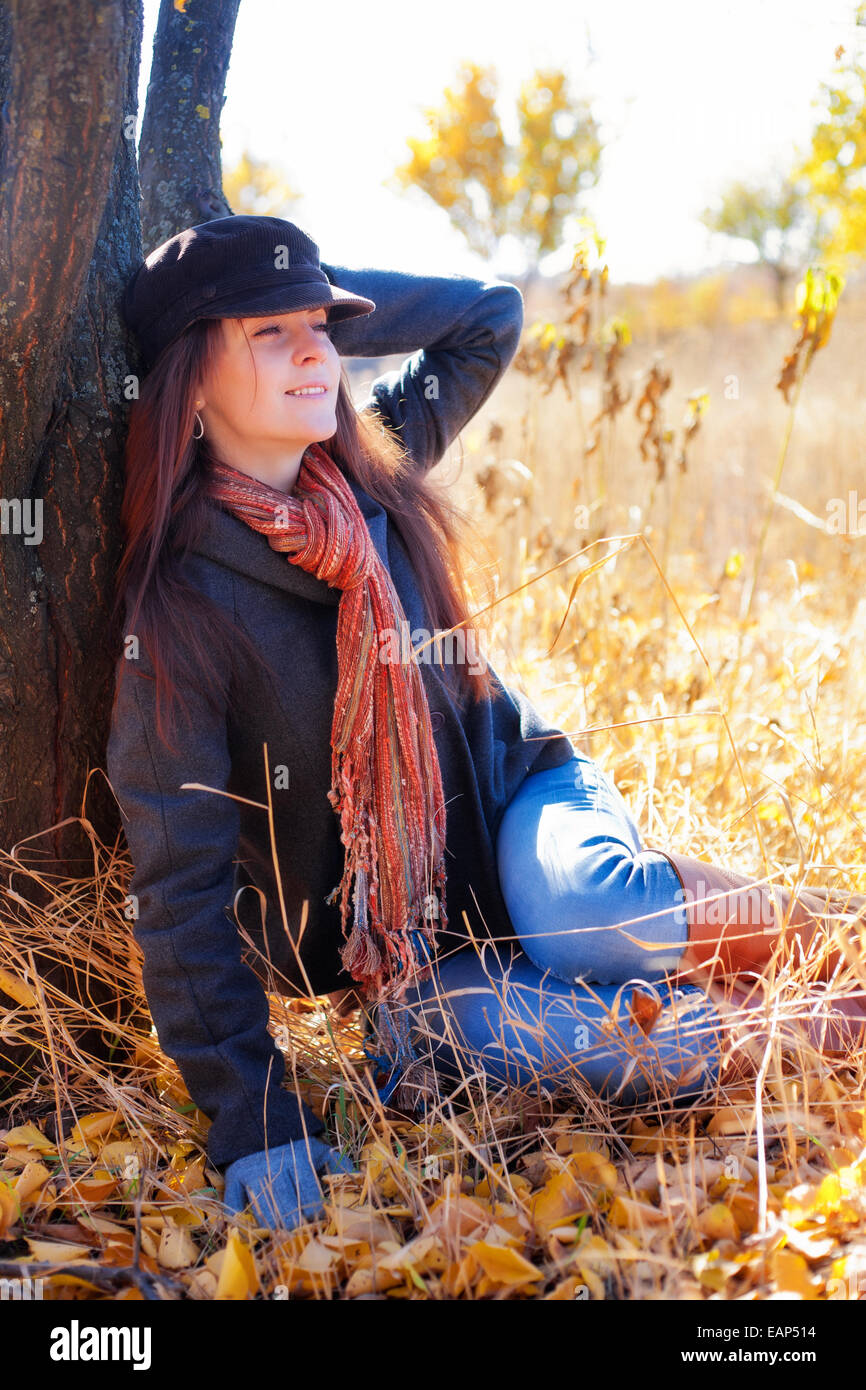 Mädchen mit Lächeln sitzt bei einem Baum an einem sonnigen Tag im Herbst Stockfoto