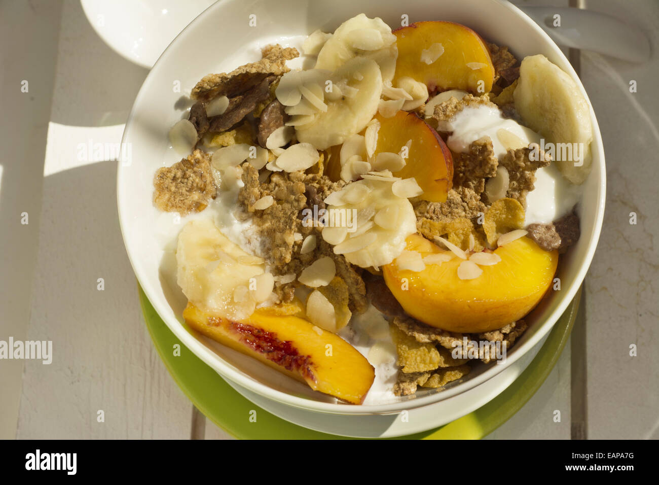 Frühstück in der Sonne, Müslischale, Pfirsich und Banane, Joghurt auf grüne Platte und warme orange Farbe. Weiß auf Holz Stockfoto