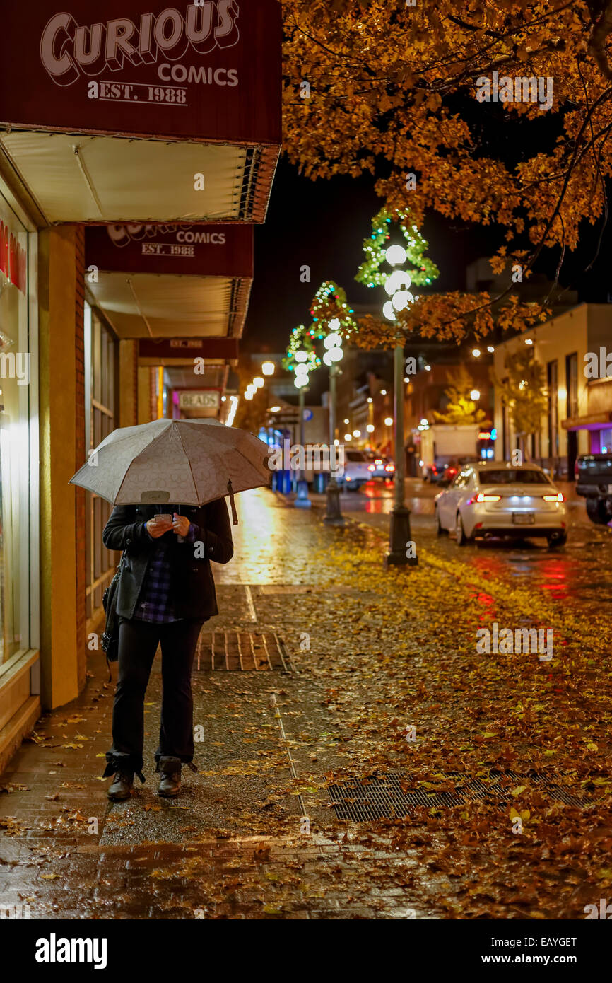 Frau am Handy mit Regenschirm auf Straße der Innenstadt am regnerischen Nacht-Victoria, British Columbia, Kanada. Stockfoto