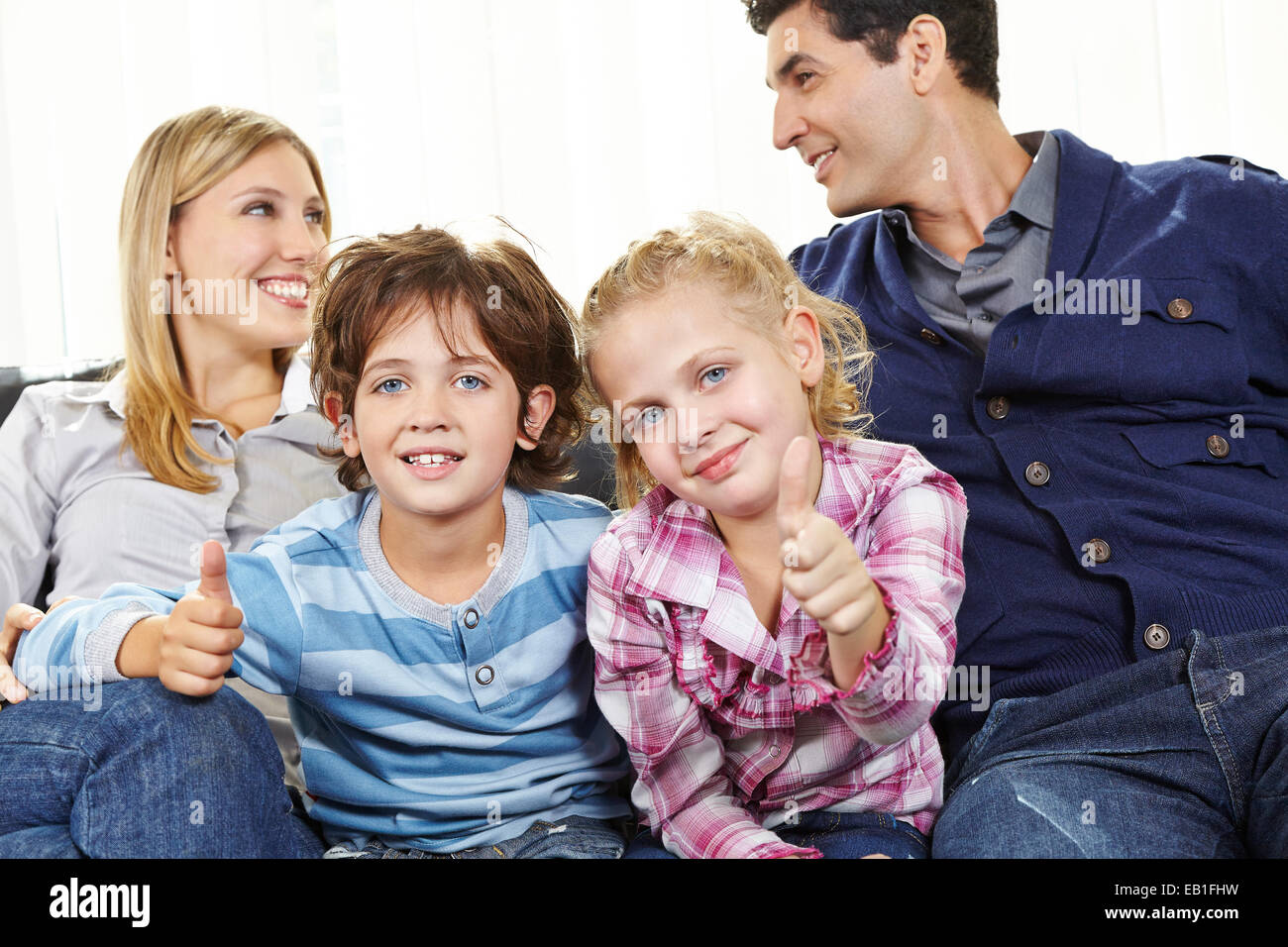 Zwei glückliche Kinder, die Daumen hochhalten, zwischen den Eltern auf dem sofa Stockfoto