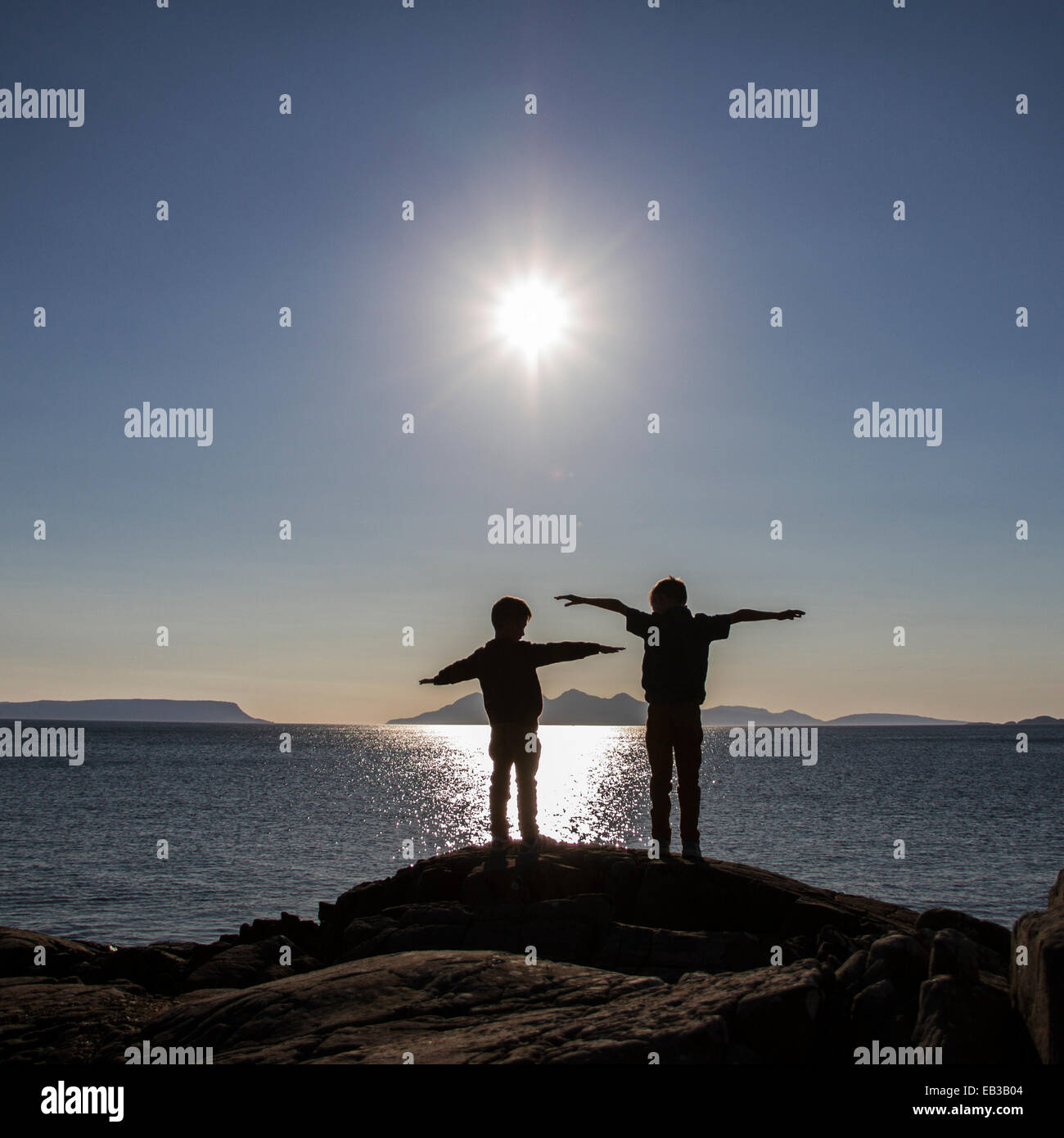 Silhouette von zwei Jungen, die mit ausgestreckten Armen auf Felsen stehen, England, Großbritannien Stockfoto