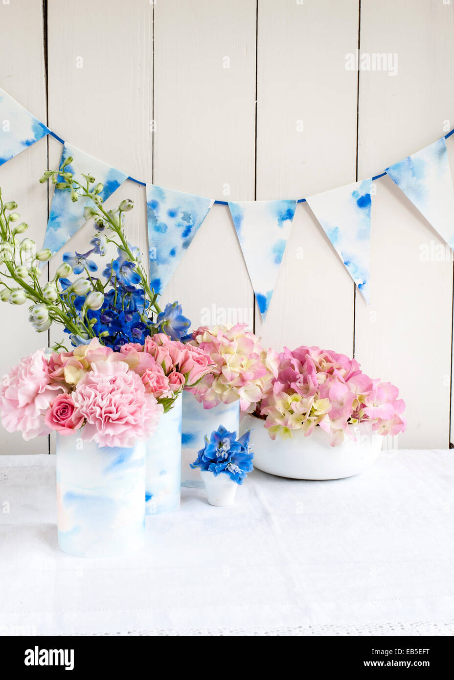 Blauer Rittersporn, rosa Rosen, Hortensien, Nelke in Dosen verziert mit bemalten Aquarellpapier, Papier-Girlanden, Stoff Stockfoto
