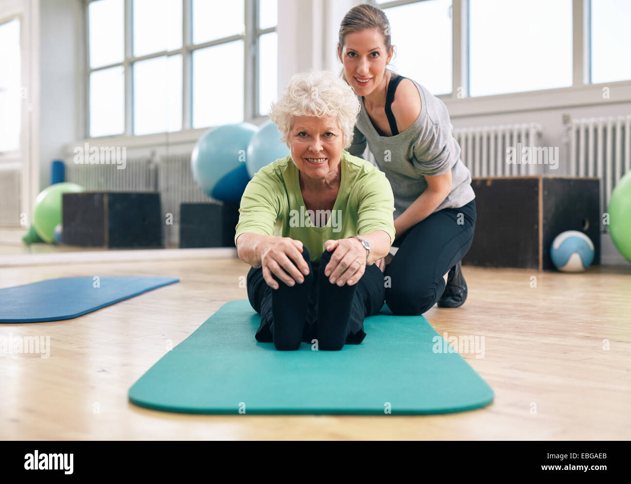 Ältere Frau, die auf der Trainingsmatte sitzt, sich nach vorne beugt und ihre Zehen mit ihrem persönlichen Trainer berührt. Fitnesstraining Stockfoto
