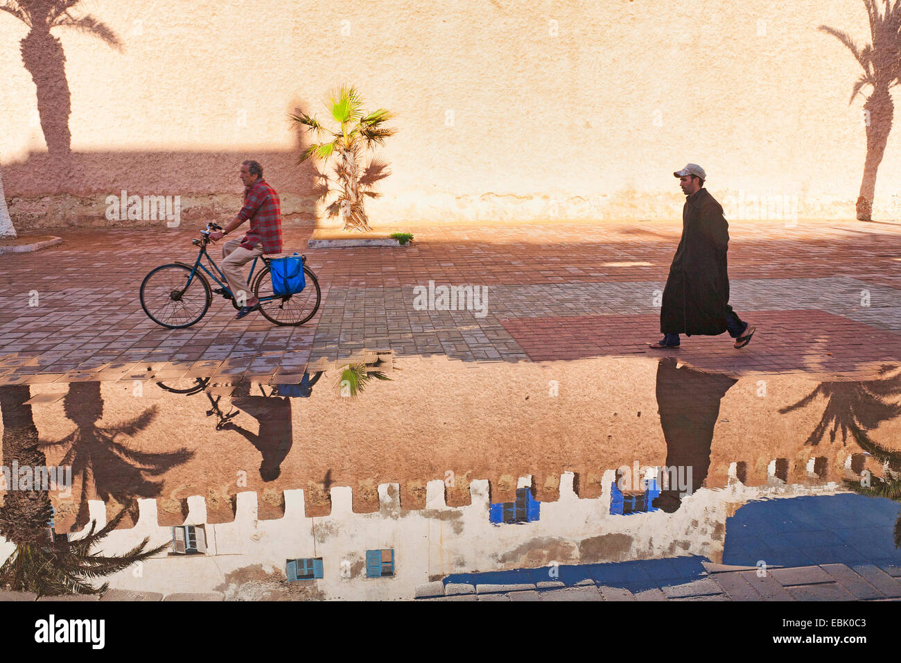 Radfahrer, Fußgänger, eine historische Stadt Wand reflektiert in Pfütze Wasser nach Ain Dusche, Marokko Essaouira Stockfoto