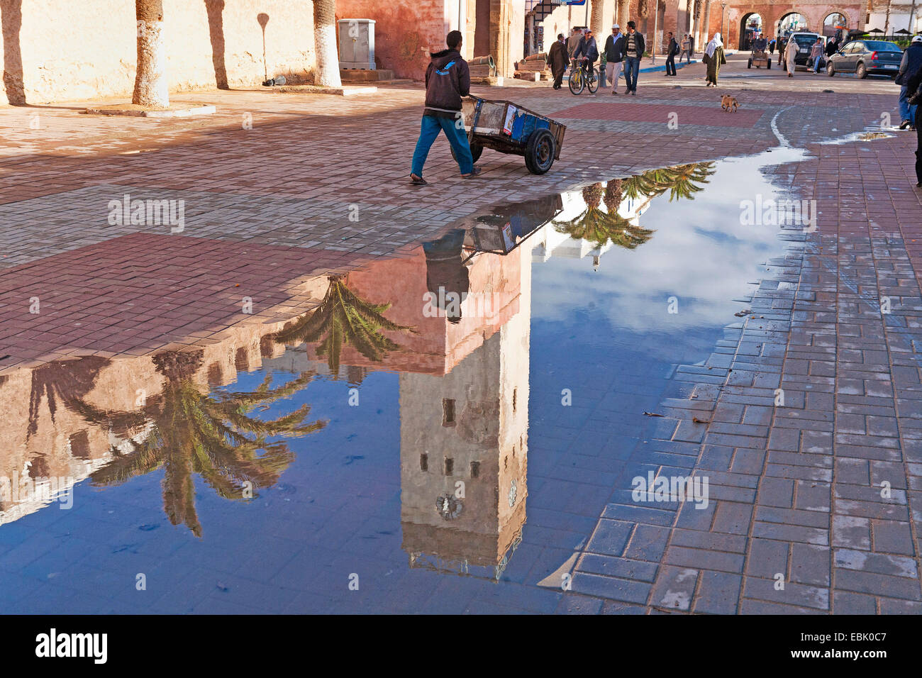 Mann mit einem Karren an der historischen Stadtmauer widerspiegelt in Pfütze Wasser nach einem Regenschauer, Marokko Essaouira Stockfoto