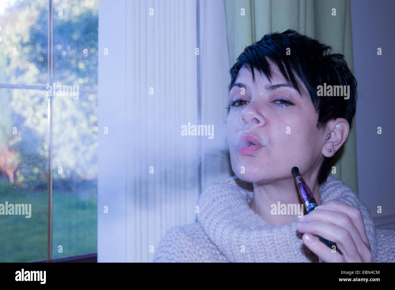 Sind e-Zigaretten cool? Schöne, kurze dunkelhaarige Frau vaping und blasen Rauch in die Kamera saß am Fenster nachdenklich Stockfoto