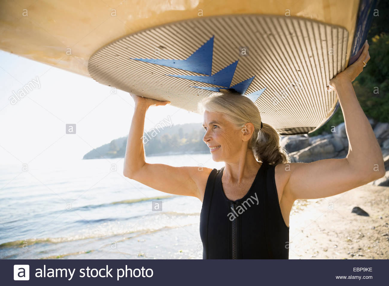 Ältere Frau hält Paddle Board overhead am Strand Stockfoto