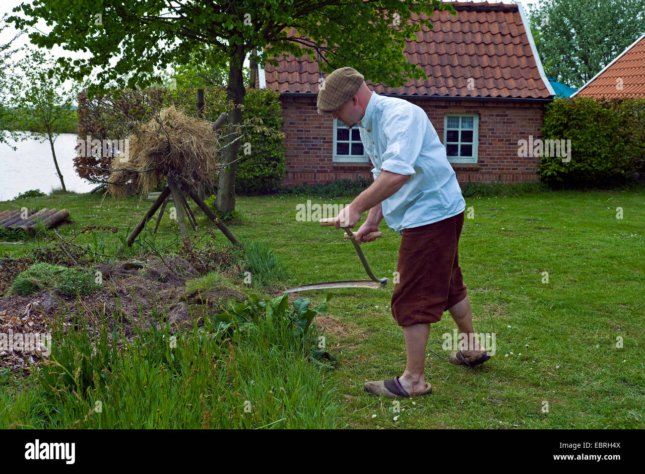 Mann in Knickerbocker, Rasen zu mähen mit einer Sense in einem Garten, Deutschland, Niedersachsen, Westgrossefehn Stockfoto