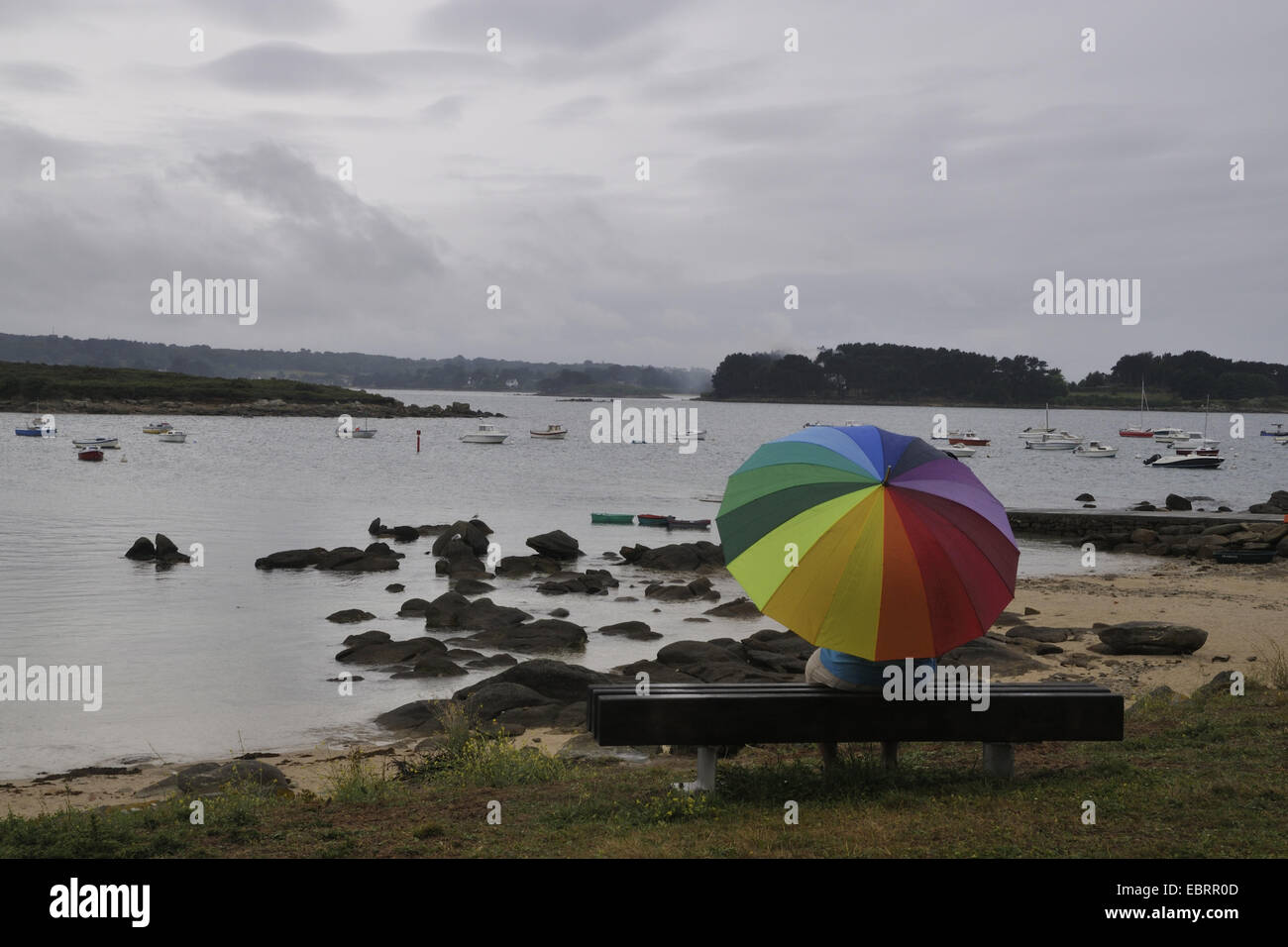 Mann im Regen mit einem bunten Schirm auf einer Bank sitzen und beobachtete das Meer, verregneten Urlaub Frankreich, Bretagne Stockfoto