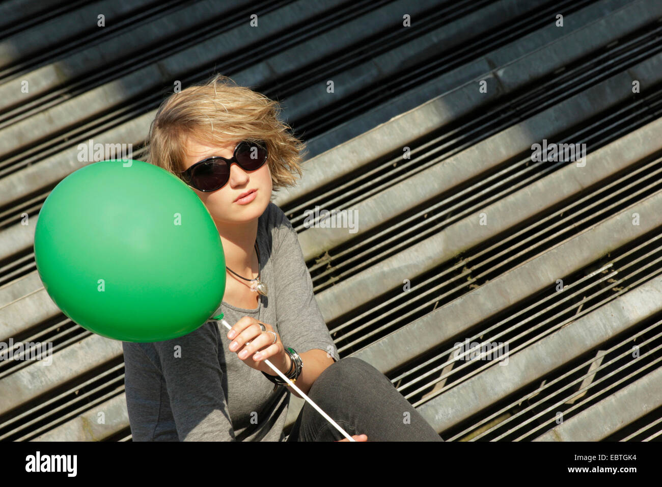 Junge, blonde Frau mit Sonnenbrille und grüner Ballon sitzt auf der Treppe, Deutschland Stockfoto