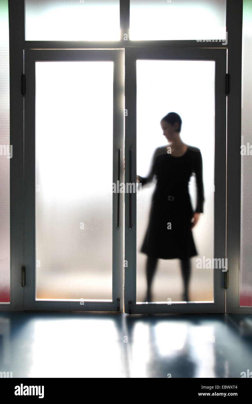 Blick durch eine Glastür in die Silhouette einer jungen Frau, die eines der beiden Türflügel geöffnet wird Stockfoto