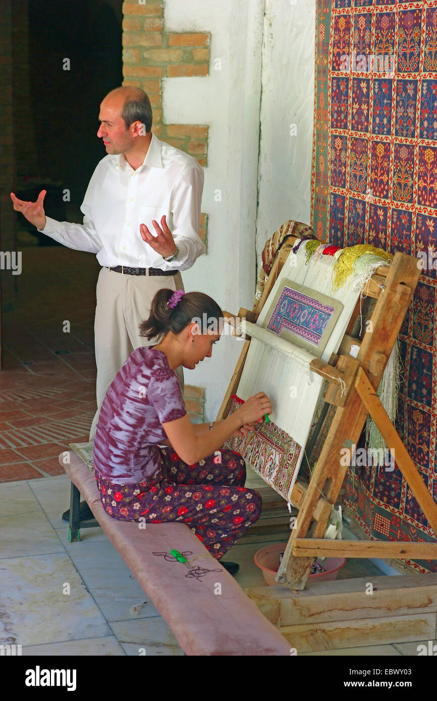 junge Frau sitzen bei der Arbeit in einem Teppich-Workshop mit einem älteren Mann stehend neben ihrem sprechen und Gestikulieren, Türkei, West-Anatolien Stockfoto