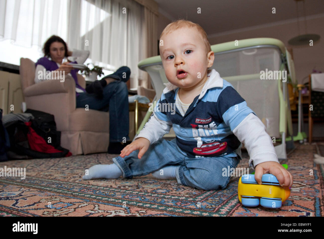 kleiner Junge spielt auf dem Boden, seine Mutter sitzt auf einem Sofa, ein Auge auf ihn Stockfoto