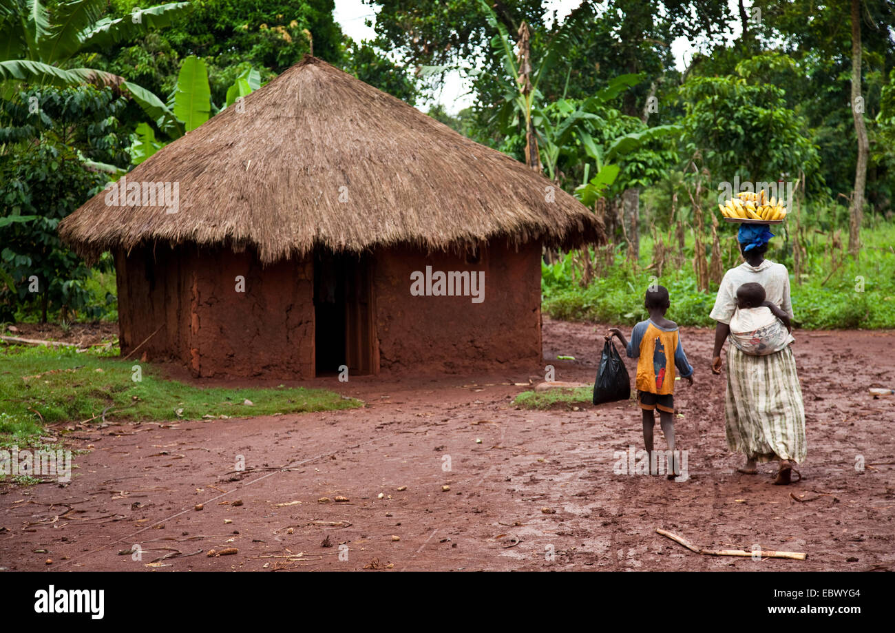 Mutter, die Transport von Bananen an den lokalen Markt mit ihr Kinder und übergibt ein Lehmhaus, Uganda, Jinja Stockfoto