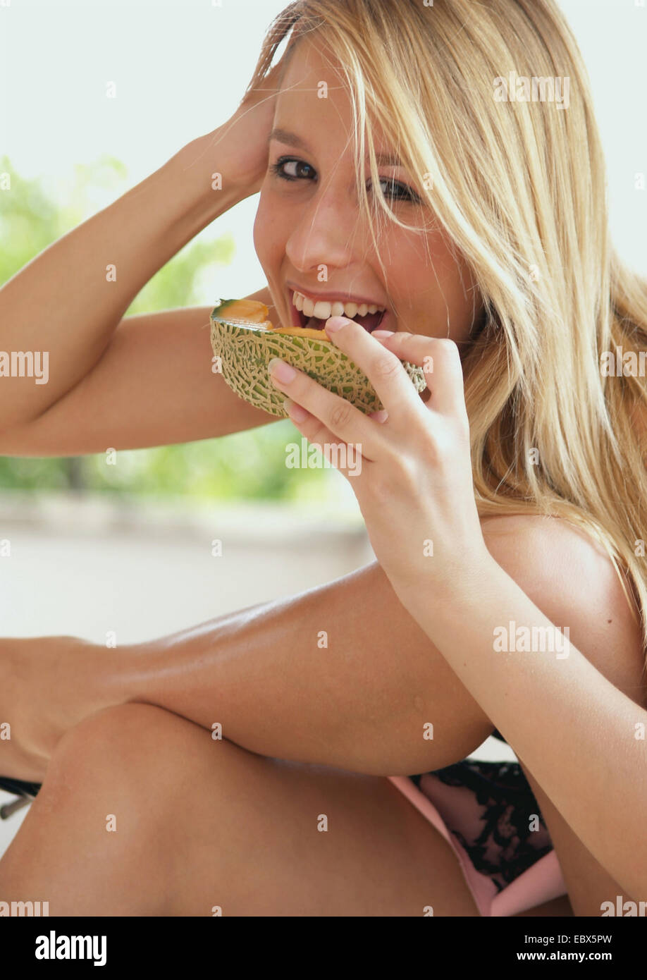 langhaarige blonde junge Frau in einem Sommer-Tuch und high Heels Essen eine Galia Melone mit einem Lächeln Stockfoto