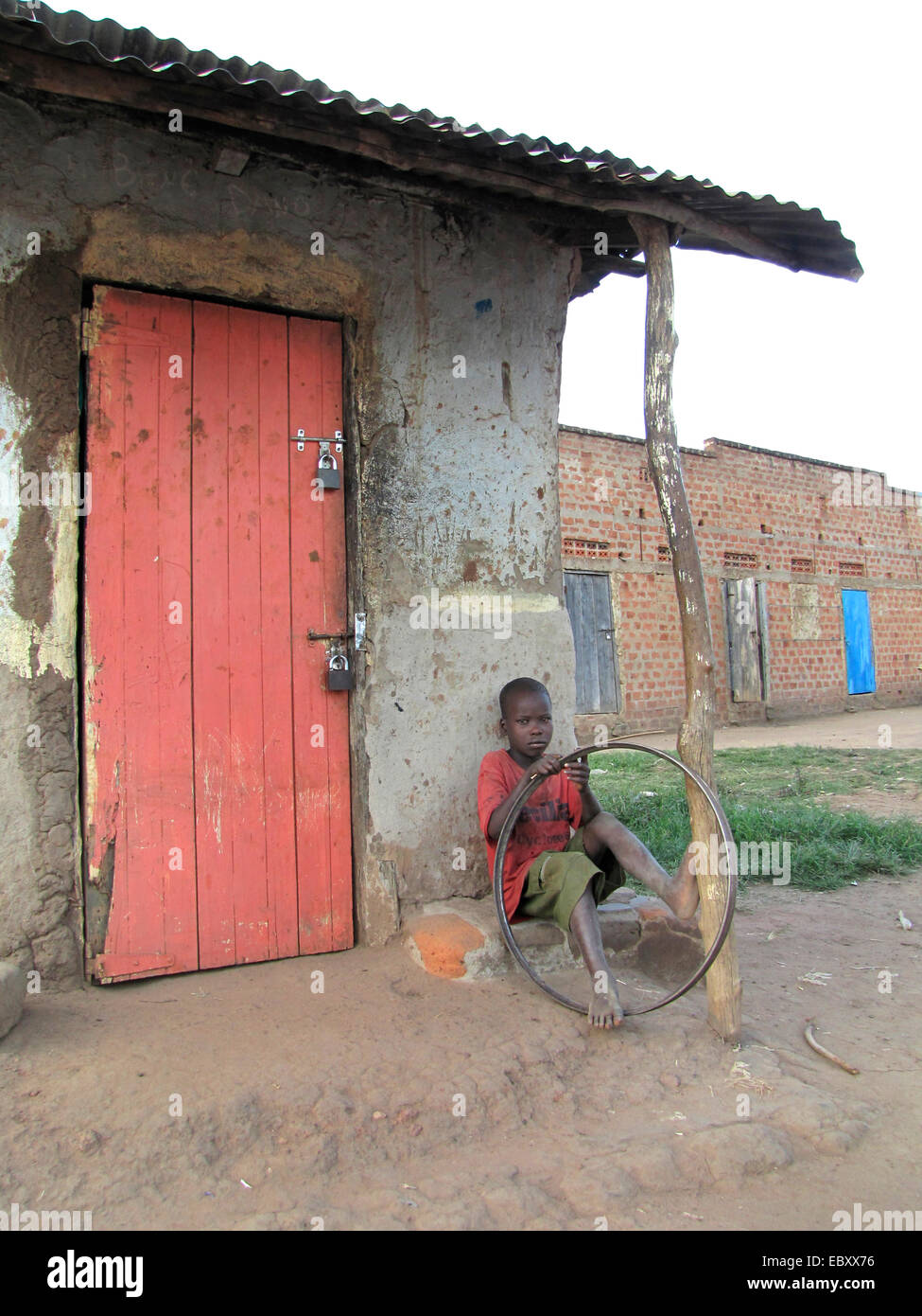 Junge sitzt neben der verschlossenen Tür ein kleines Geschäft mit einem alten Fahrradreifen in Händen, Gulu, Uganda, Gulu Stockfoto