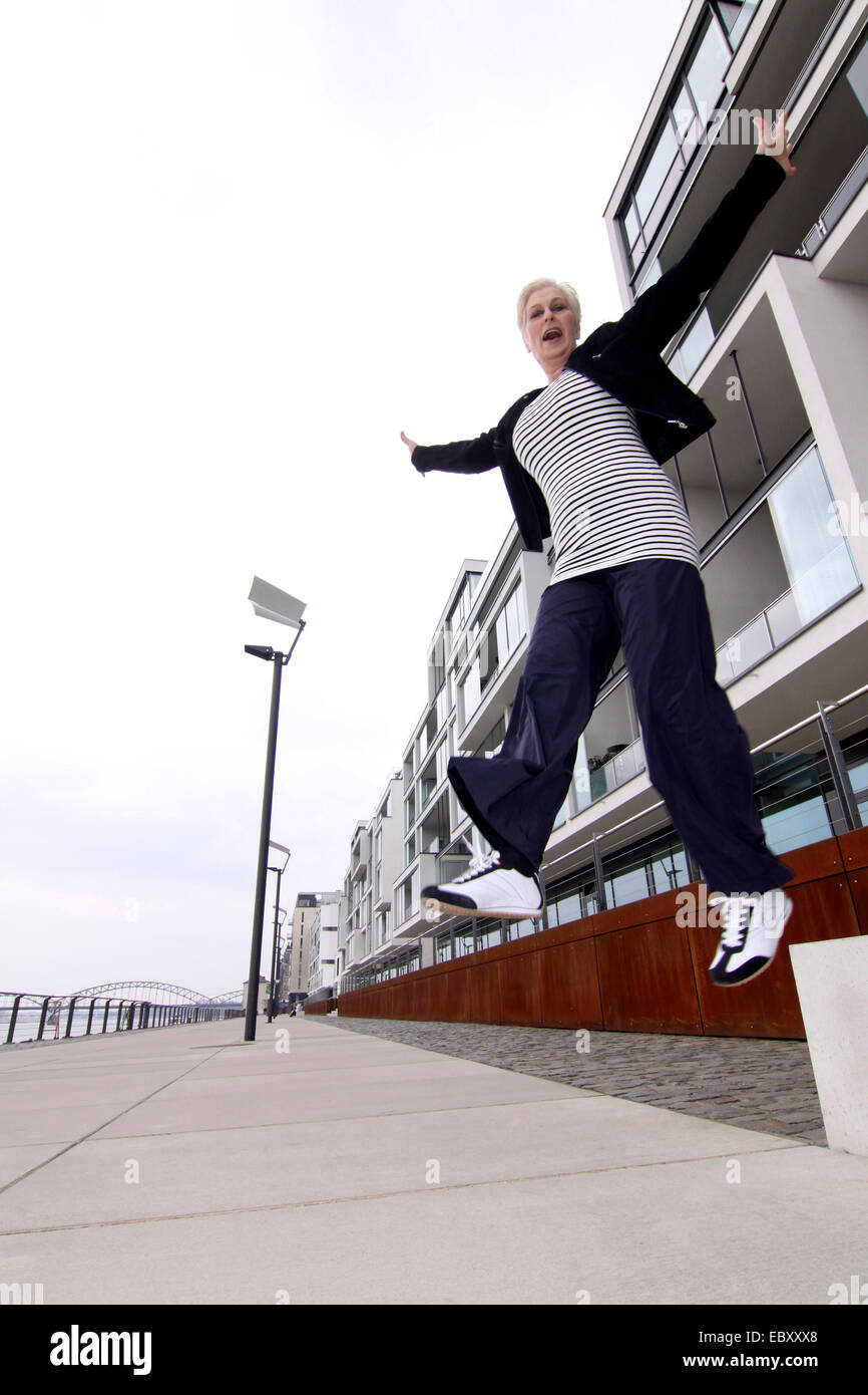 attraktive weißhaarige Frau in Freizeitkleidung in die Luft sprang an einem Rheinpromenade, Deutschland, Nordrhein-Westfalen, Agrippinawerft, Köln Stockfoto