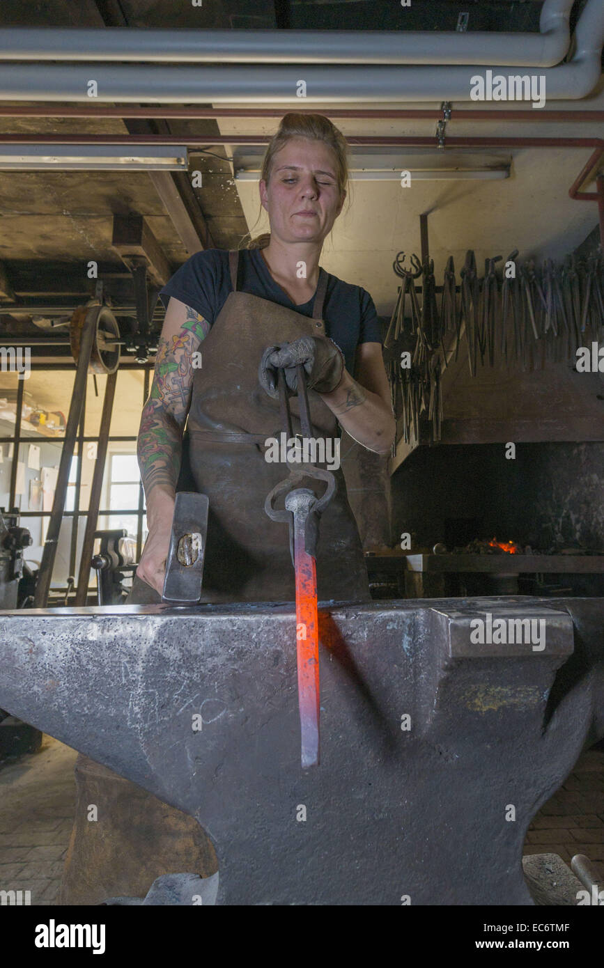 weiblichen Schmied Inspektion ein Redhot Stück Stahl am Amboss, Fenster im Hintergrund Stockfoto