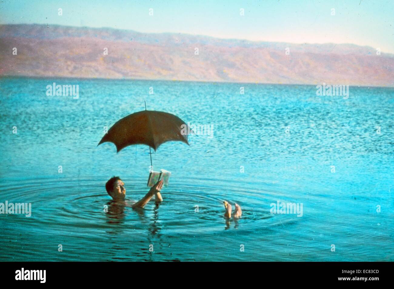 Farbfoto eines Mannes im Toten Meer schweben, während ein Buch zu lesen und einen Regenschirm halten. Datiert 1930 Stockfoto