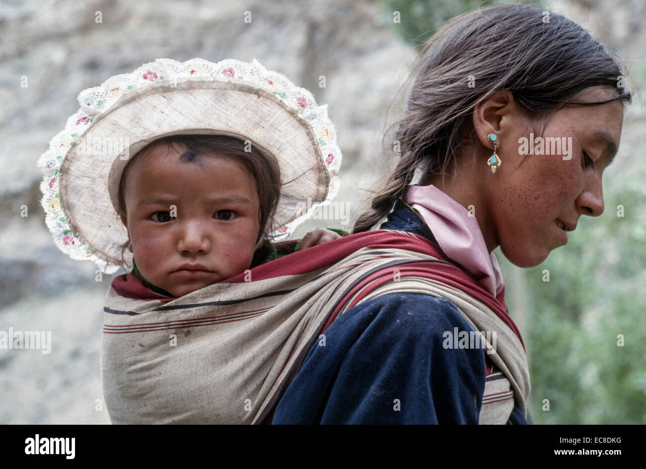 Ladakh-Mutter-Kind auf Rücken Sonne Hut getragen Tuch Papoose junge Frau dunkles Haar Kinderprofil Stockfoto