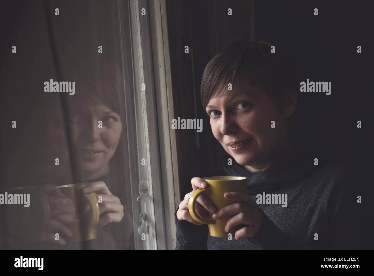 Schöne kaukasischen Erwachsenen lächelnde Frau trinken und genießen Kaffee im dunklen Raum durch das Fenster. Stockfoto