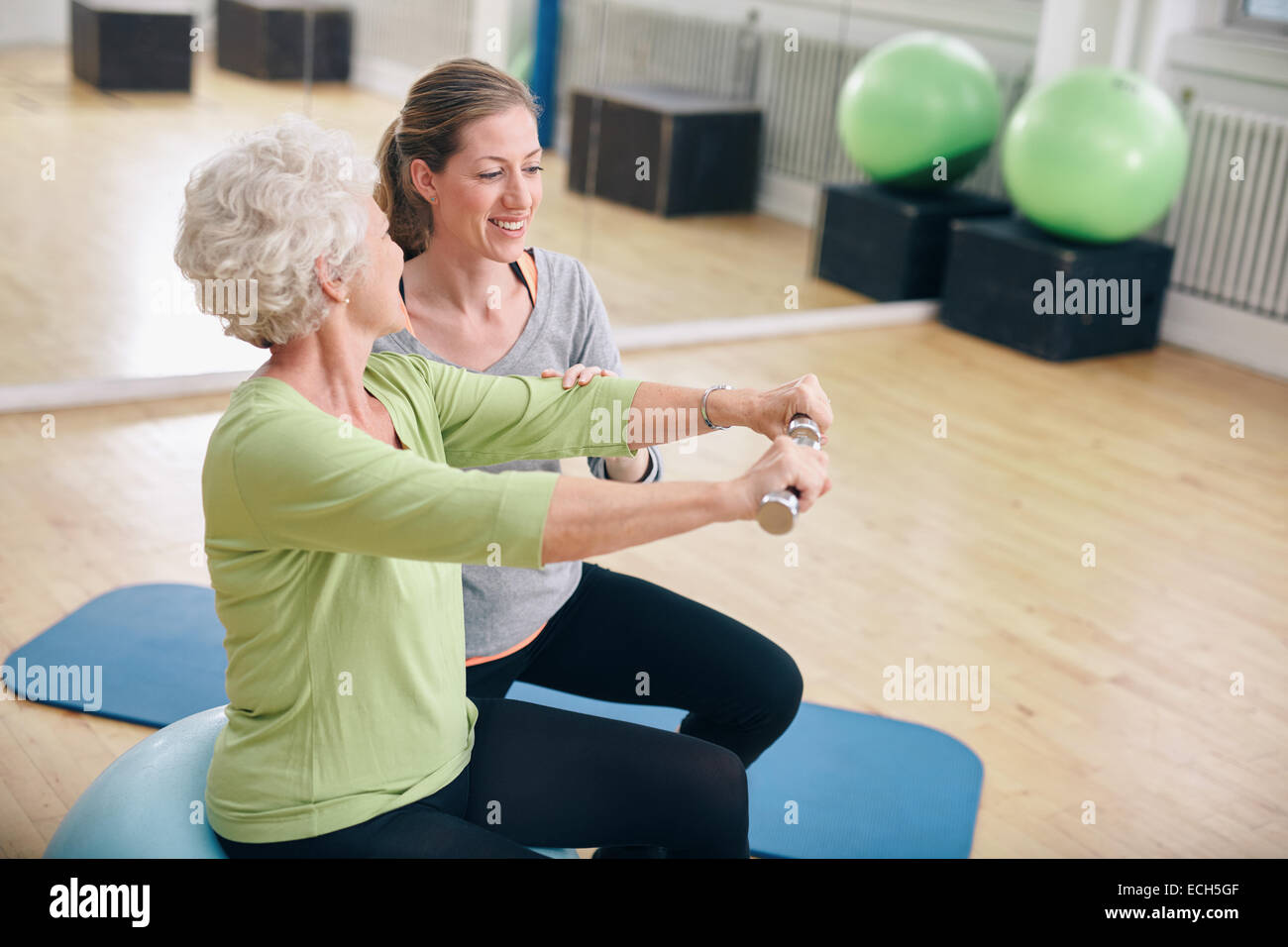 Ältere Frau Training mit Gewichten in der Turnhalle von einem jungen weiblichen Trainer unterstützt. Alte Frau Aufhebung Hanteln mit Hilfe. Stockfoto