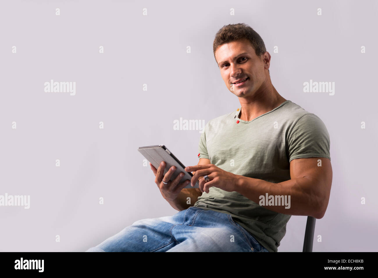 Muskulösen jungen Mann sitzend auf Stuhl Lesen von Ebook Gerät, lächelnd in die Kamera Stockfoto