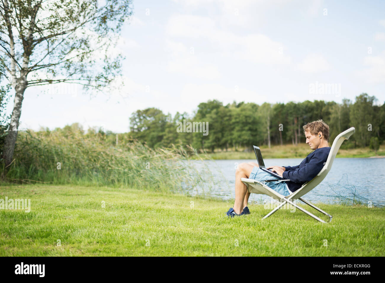 Voller Länge Seitenansicht von reifer Mann mit Laptop auf Liegestuhl am Seeufer Stockfoto