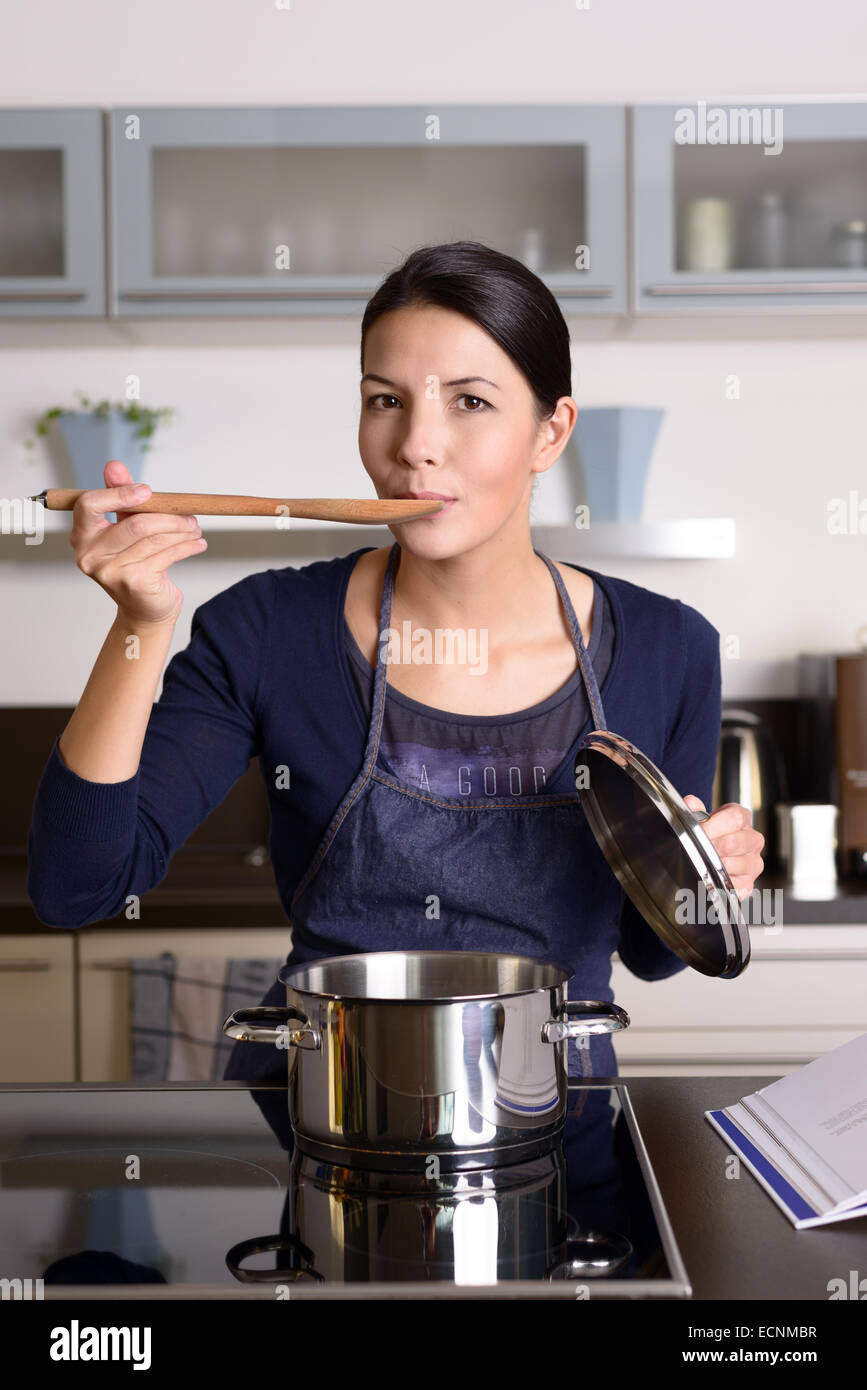 Junge Hausfrau, die ihre Küche wie sie das Abendessen in ihrer Küche Probenahme direkt aus dem Topf auf dem Herd mit einem w bereitet Stockfoto