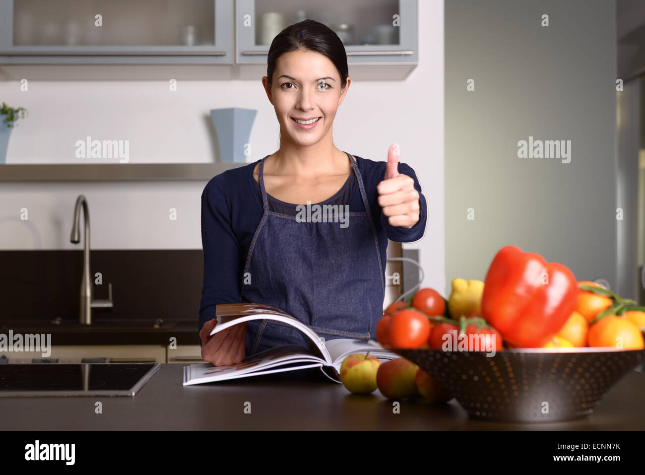 Glücklich im mittleren Alter Frau tragen Schürze stehen an der Küchentheke mit Rezeptbuch, ein positives Daumen aufgeben Stockfoto