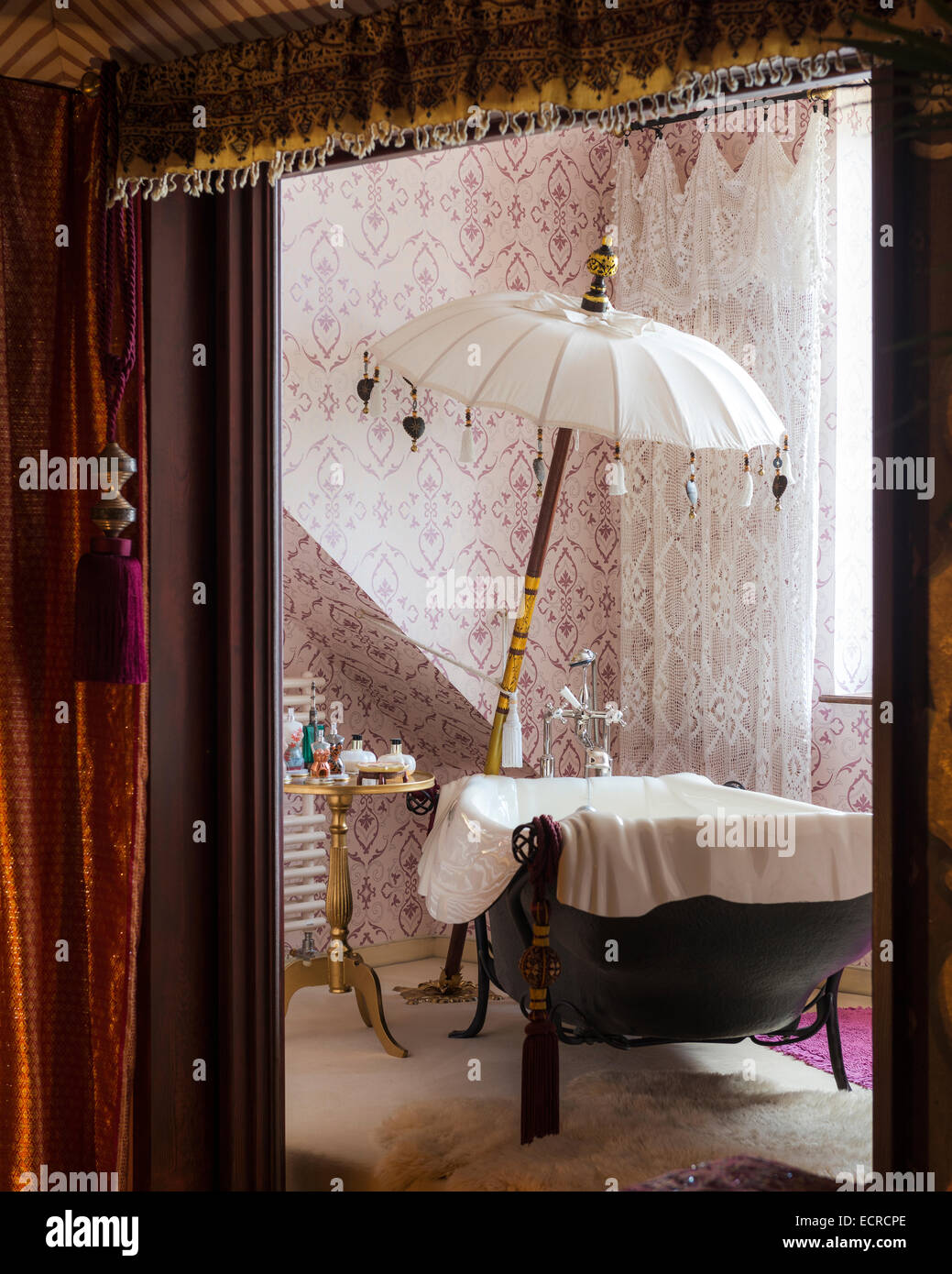 Freistehende Badewanne mit Sonnenschirm im Badezimmer mit gemusterten Tapeten und Gardinen Stockfoto