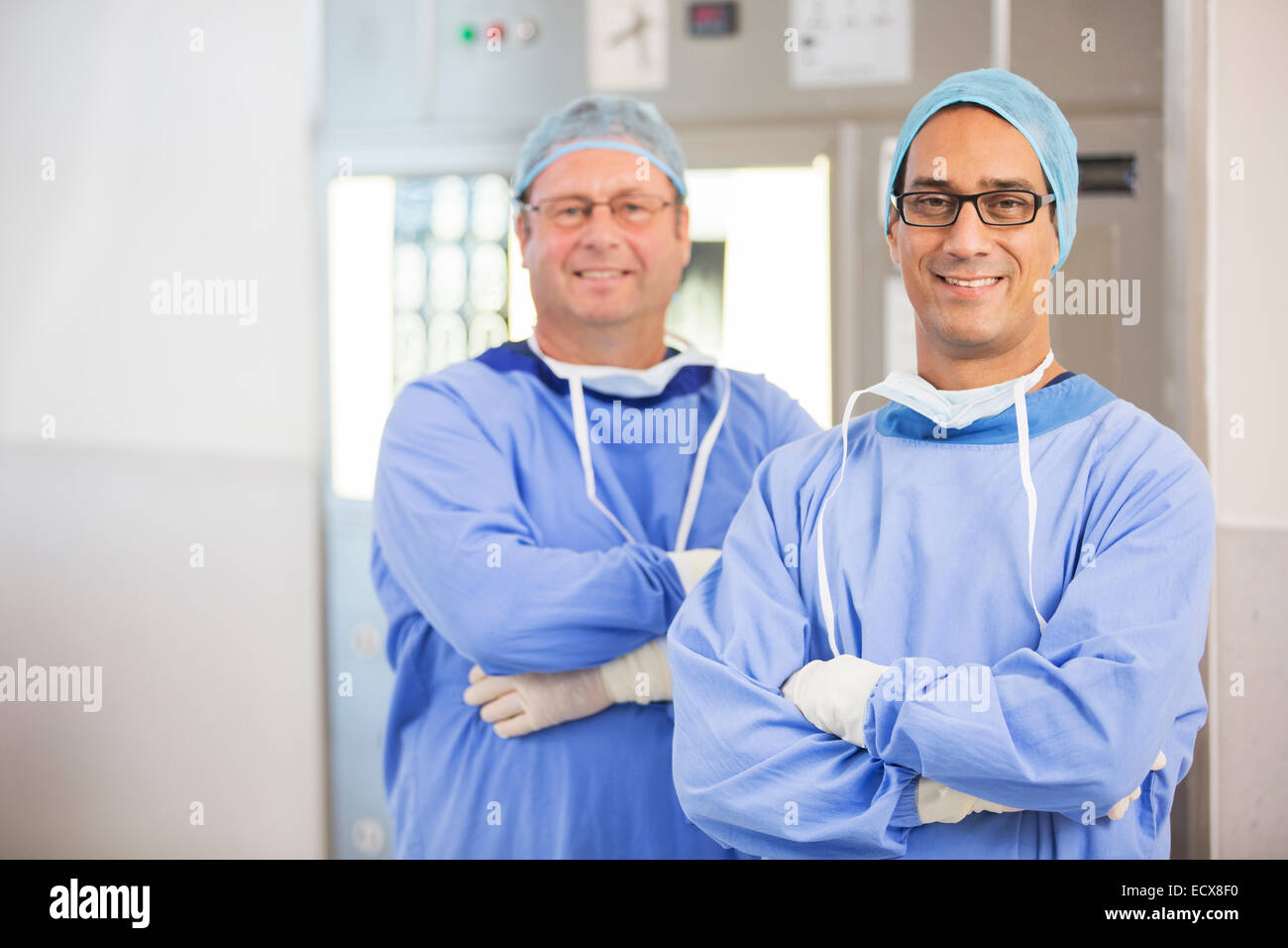 Zwei lächelnde Ärzte mit verschränkten Armen und tragen von OP-Kleidung und Brillen im Krankenhaus Stockfoto