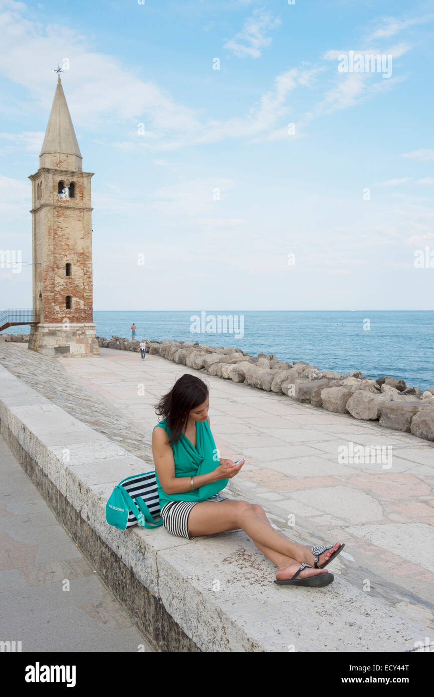 Frau mit Handy sitzt an der Wand vor der Kirche Madonna Dell ' Angelo, Adria, Caorle, Veneto Region, Italien Stockfoto
