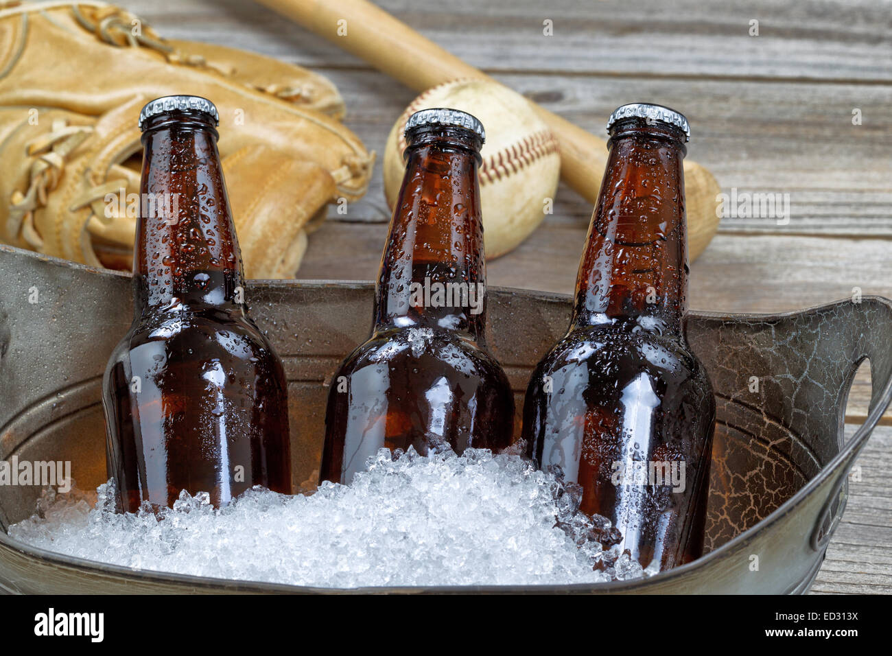 Schließen sich drei braune Kälte Flaschenbiere, crushed Eis in Metallbehälter und Baseball-Ausrüstung im Hintergrund auf rustikalen Holz Stockfoto