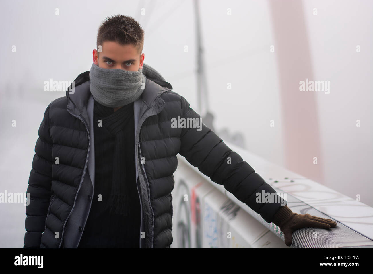 Junger Mann im Winter Mode stehen im Freien in einen warmen Mantel und Handschuhe mit seinem grauen Schal gewickelt runden die untere Hälfte des Hallo Stockfoto