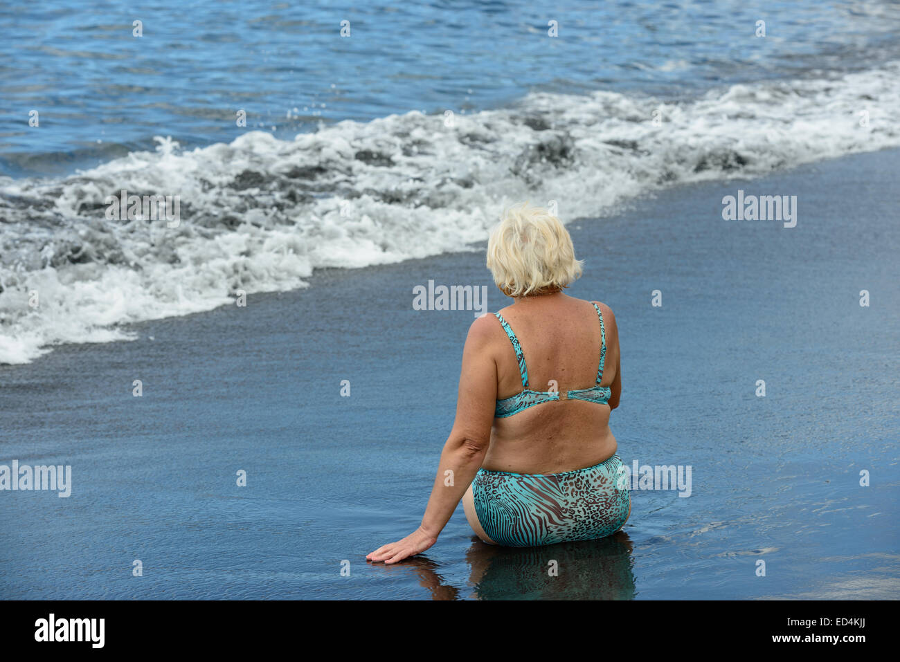 Alte Frau ist Sittig zurück zur Kamera auf Grund der Brandung am nassen schwarzen vulkanischen Sand der Arena Strand in Puerto de Santiago, Tener Stockfoto