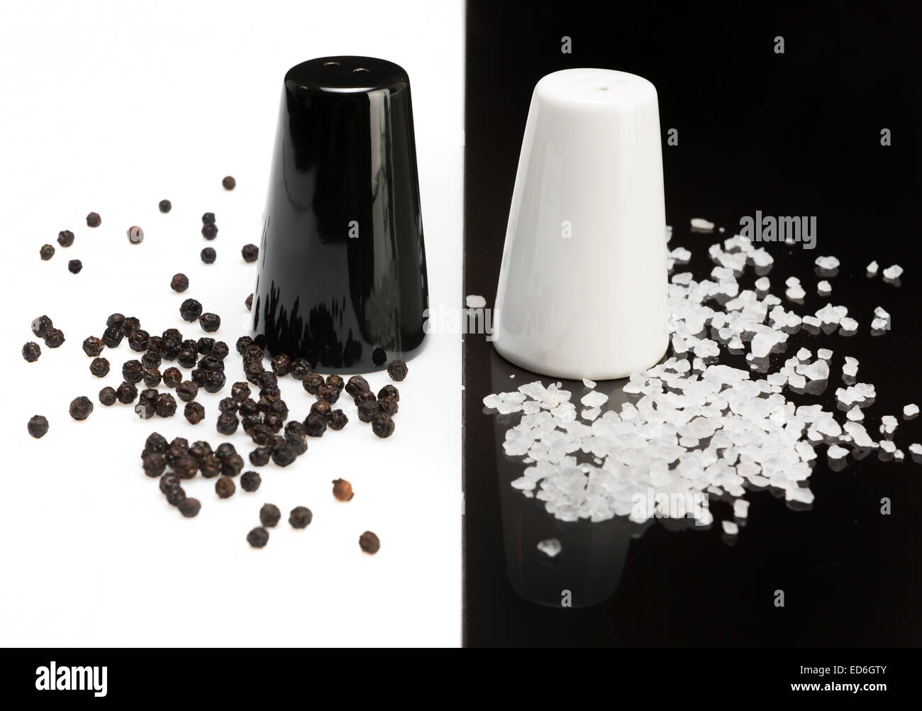 schwarzer Pfeffer Shaker auf weißem Hintergrund und Pfefferkörner, weiße Salzstreuer auf einem schwarzen Hintergrund und Salzkristalle Stockfoto