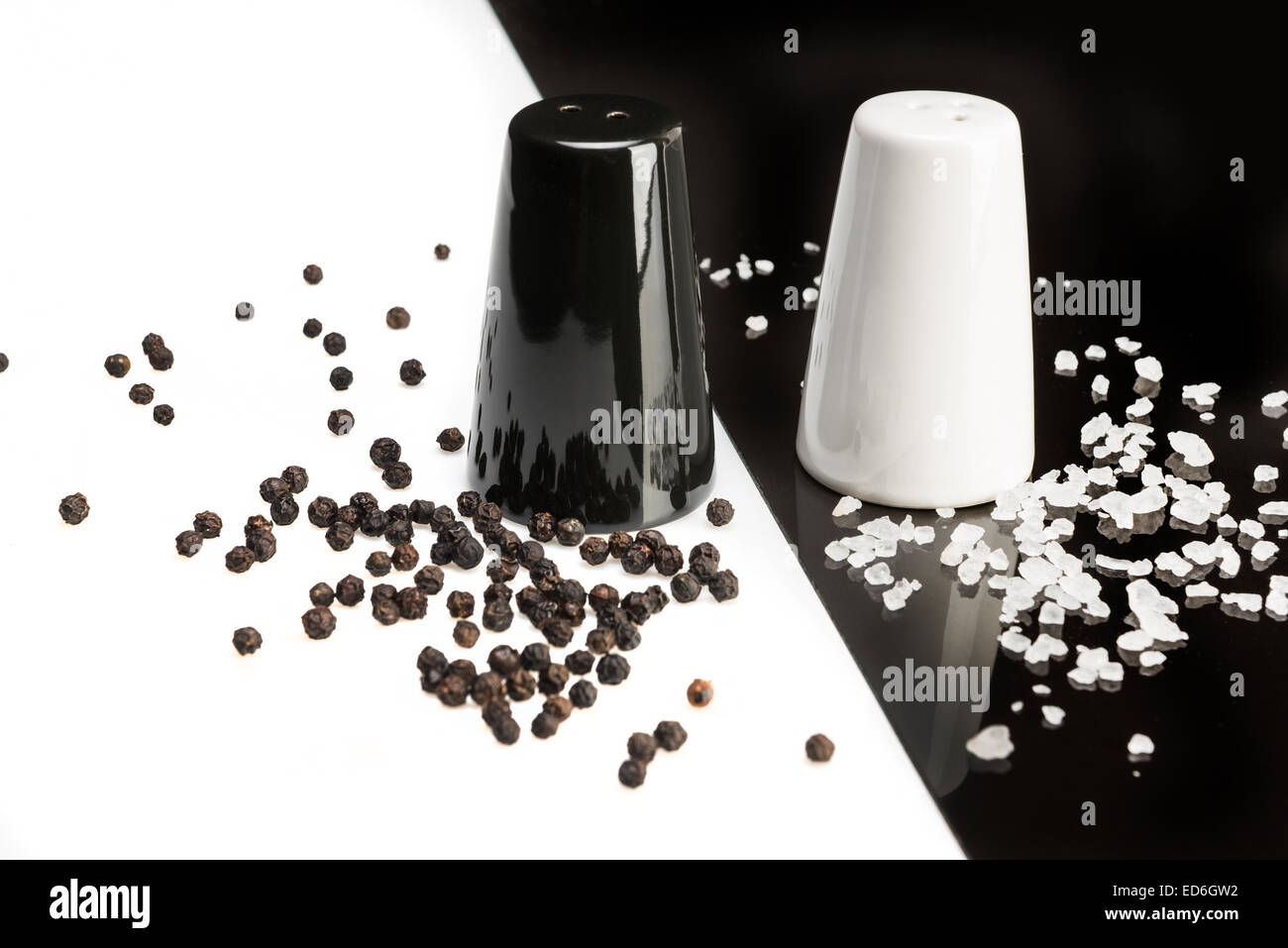schwarzer Pfeffer Shaker auf weißem Hintergrund und Pfefferkörner, weiße Salzstreuer auf einem schwarzen Hintergrund und Salzkristalle Stockfoto