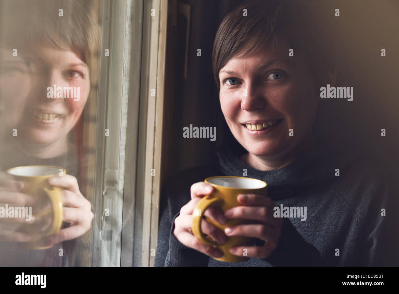 Schöne kaukasischen Erwachsenen lächelnde Frau trinken und genießen Kaffee im dunklen Raum durch das Fenster. Selektiven Fokus mit sha Stockfoto